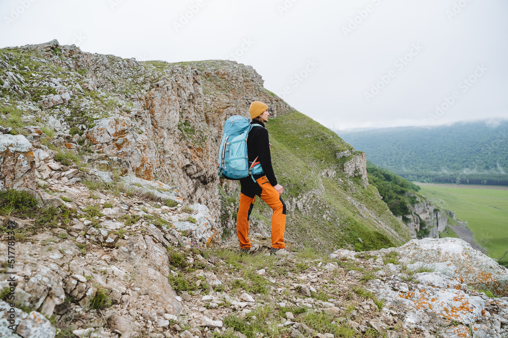 A man's behind view in the mountains with a backpack the traveler stands on the edge of a cliff a guy admires the view of nature beautiful mountain scenery summer vacation
