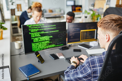 Rear view of young IT specialist writing codes of software sitting at office desk in front of computer monitors