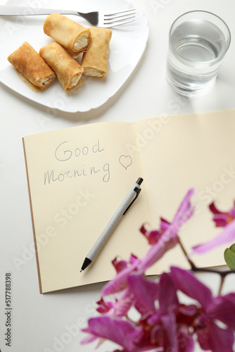 Notebook with inscription Good Morning, stuffed crepes and glass of water near beautiful blooming orchid on white table, above view