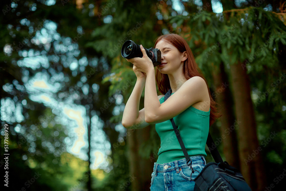 Young woman with ginger hair enjoys nature and takes photos. Pretty girl wearing green top holds the camera.