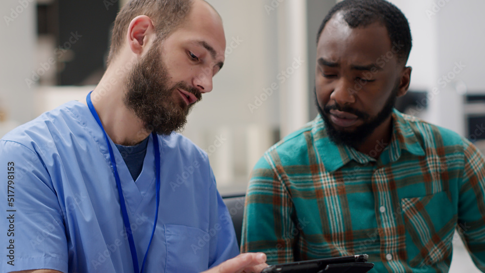 Fototapeta premium African american man and male nurse looking at digital tablet to find healthcare diagnosis and recovery treatment in waiting room lobby. People doing examination at medical clinic.