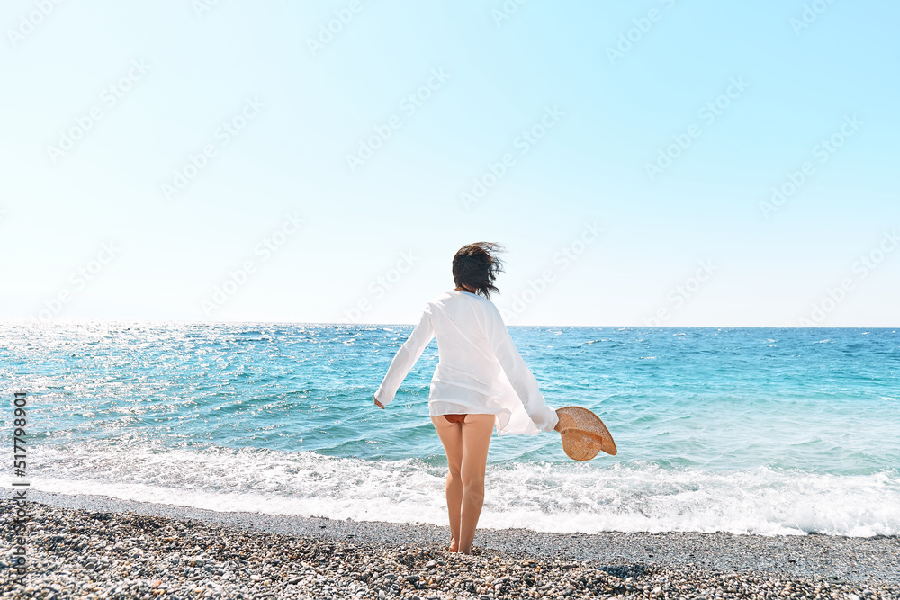 Back view of carefree woman in straw hat and white shirt walking on pebble beach. Relaxing and enjoy holiday at the sea in summertime. Unity with nature. Wellness, success, freedom and travel concept Back view of carefree woman in straw hat and white shirt walking on pebble beach. Relaxing and enjoy holiday at the sea in summertime. Unity with nature. Wellness, success, freedom and travel concept
