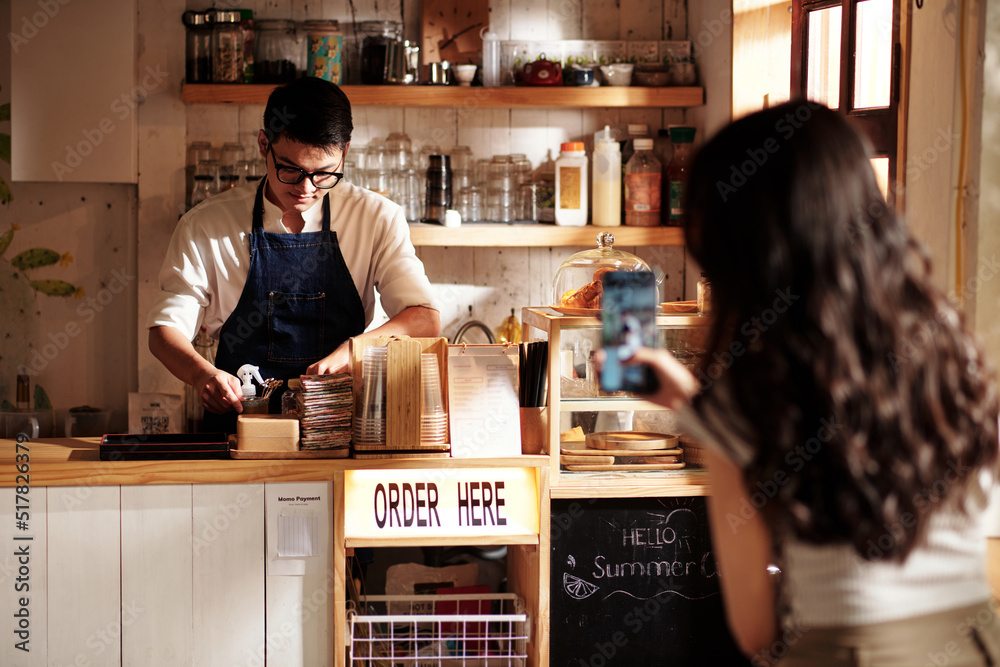 Young woman taking photo of working barista for social media account of coffeeshop