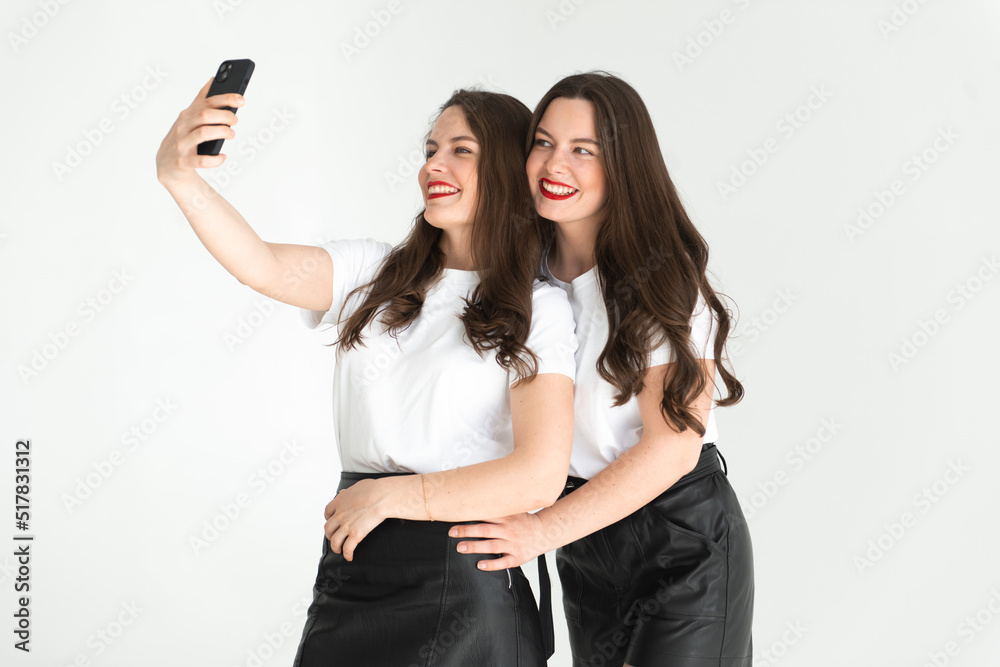 Two beautiful women twin sisters having fun together, posing white background and tasing selfie Two beautiful women twin sisters having fun together, posing white background and tasing selfie