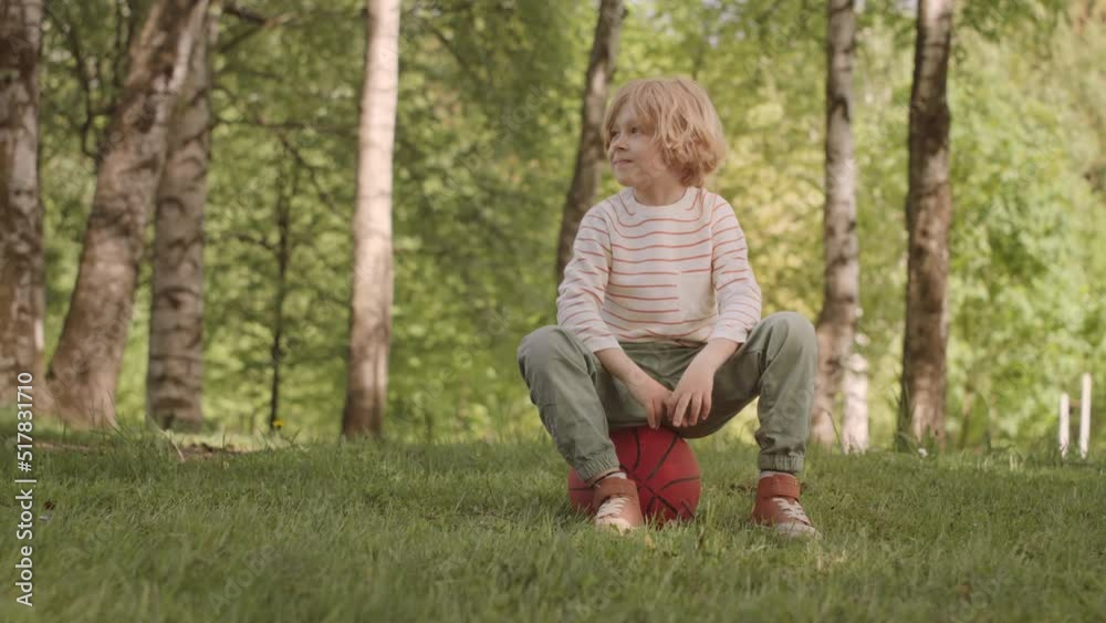 Slowmo portrait of cute blonde 7 year old boy sitting on basketball on green grass in park smiling at camera Slowmo portrait of cute blonde 7 year old boy sitting on basketball on green grass in park smiling at camera