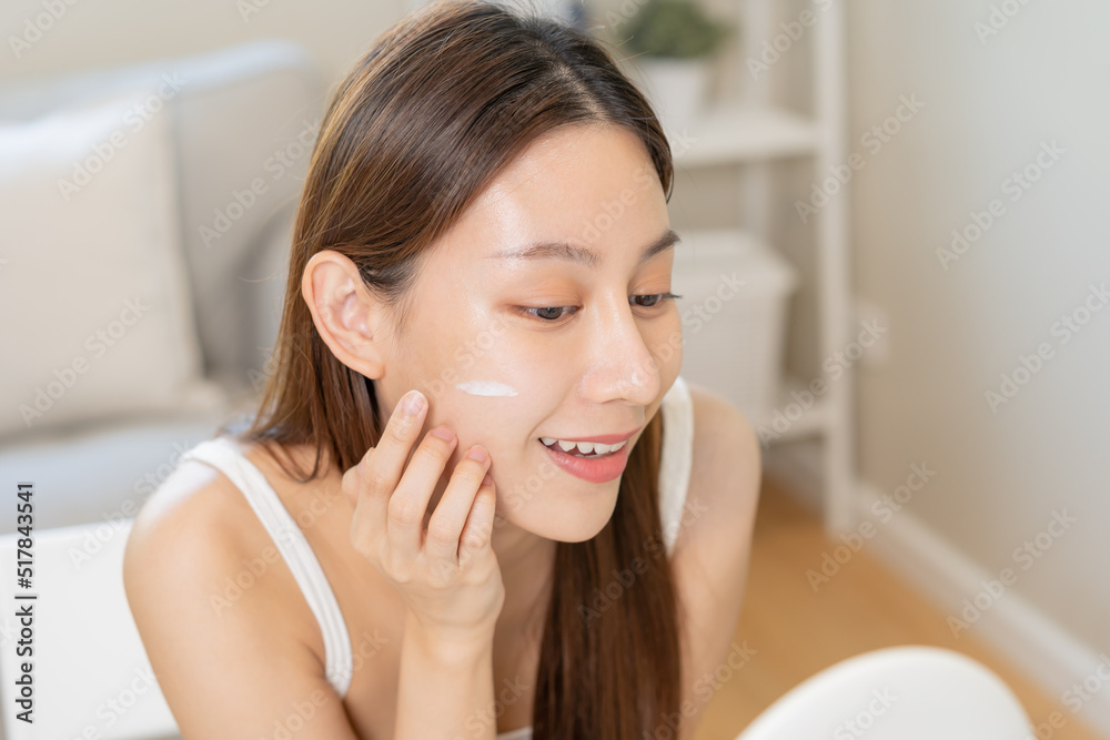 Fresh healthy skin, beautiful smile of asian young woman, girl looking at mirror, applying moisturizer on her face, putting cream treatment before makeup cosmetic routine at home. Facial Beauty. Fresh healthy skin, beautiful smile of asian young woman, girl looking at mirror, applying moisturizer on her face, putting cream treatment before makeup cosmetic routine at home. Facial Beauty.