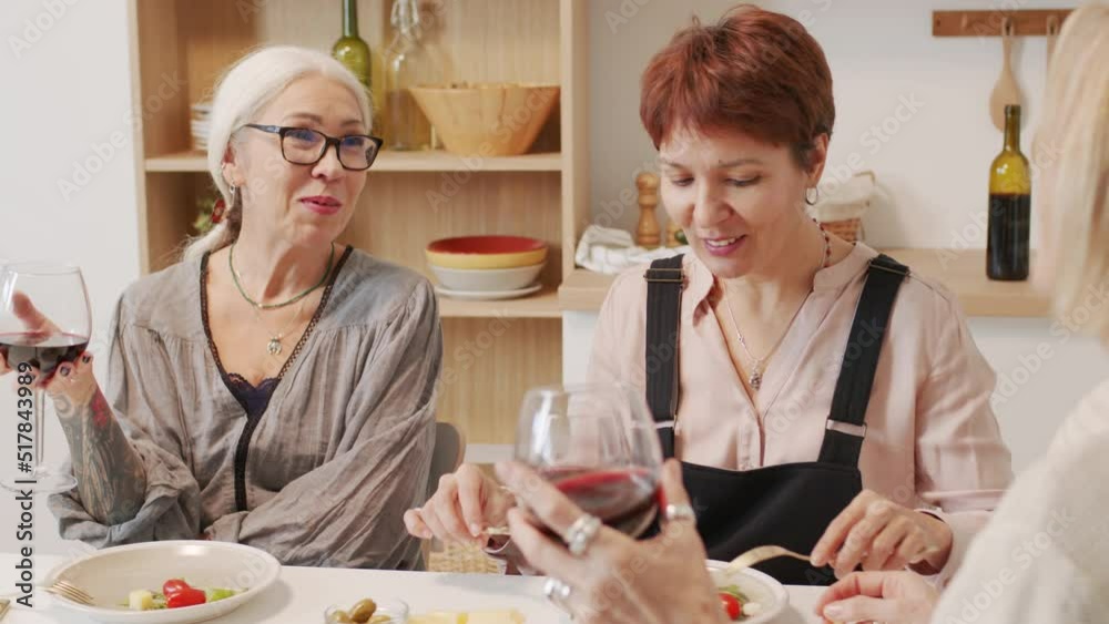 Three female best friends talking and clinking glasses of red wine while having dinner together at table in minimalist kitchen Three female best friends talking and clinking glasses of red wine while having dinner together at table in minimalist kitchen