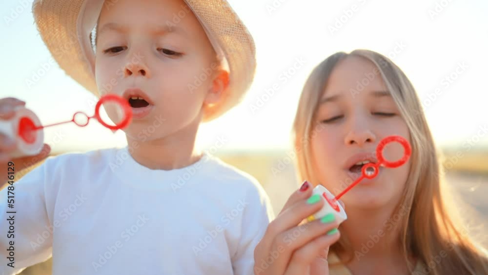 Teenage sister stands and smiles next to little brother who blows and gets soap bubbles in outdoor in field. Family walk on weekend out of town in countryside. Leisure activity. Healthy lifestyle. Teenage sister stands and smiles next to little brother who blows and gets soap bubbles in outdoor in field. Family walk on weekend out of town in countryside. Leisure activity. Healthy lifestyle.