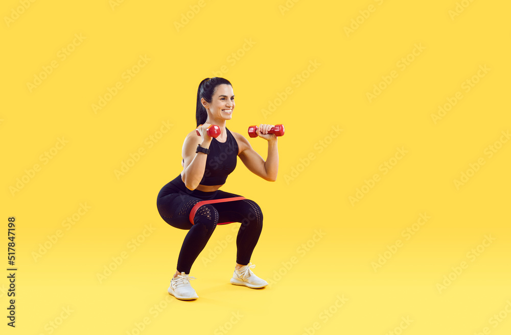 Smiling active young woman athlete or coach make squats with dumbbells and fitness bands. Happy toned female trainer workout exercise on yellow studio background. Sport lifestyle.