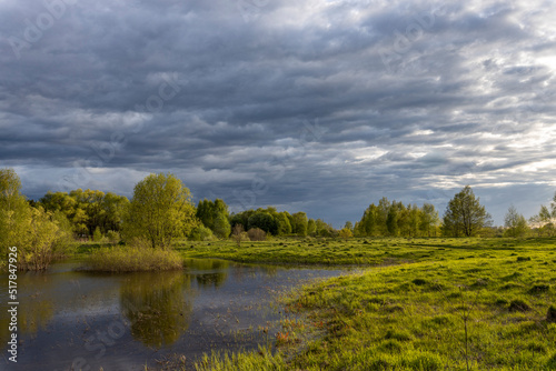 Rural landscape on a spring evening. the dramatic sky is reflected in the river. Bright sun rays illuminate the grass in the foreground.