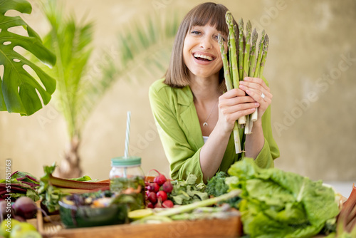 Portrait of a young cheerful woman holds a bunch of asparagus while sitting by the table full of fresh vegetables, fruits and greens. Healthy eating and lifestyle concept