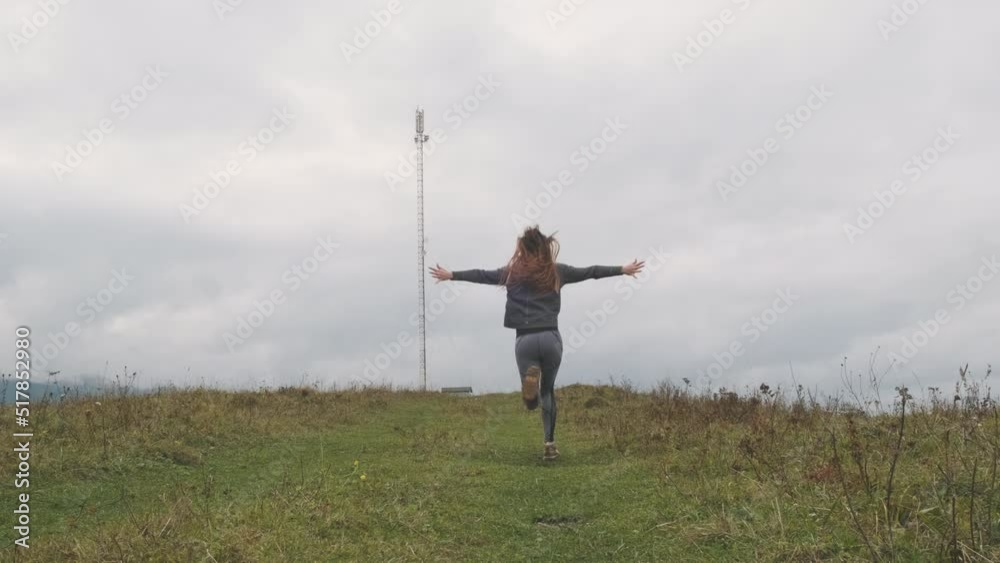 Beautiful female runs along green hill towards the clouds, spreading her arms to sides. Bottom view from behind, mountains, pillar. Concept of happiness, travel, goal achievement, joy, nature, hiking Beautiful female runs along green hill towards the clouds, spreading her arms to sides. Bottom view from behind, mountains, pillar. Concept of happiness, travel, goal achievement, joy, nature, hiking