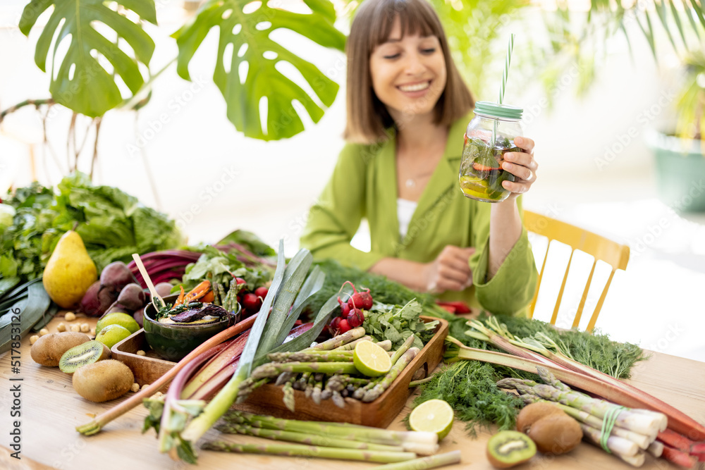 Obraz premium Young woman takes a bottle with drink while sitting by the table full of fresh vegetables, fruits and greens indoors. Concept of healthy vegan food and lifestyle