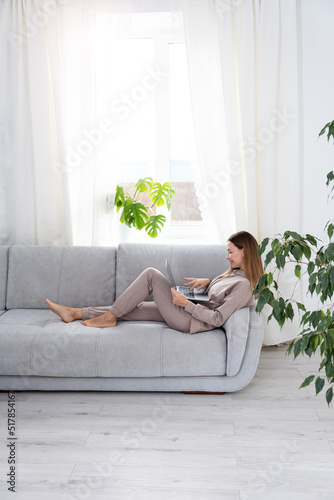 Young or middle age woman sitting with laptop on grey couch in home office with monstera plant. Concept of remote workplace and working at home.
