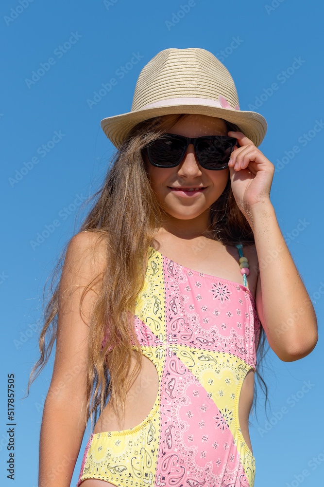 A teenage girl in a bathing suit, sunglasses and a hat against the sky.
