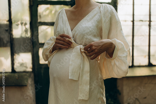 a young pregnant woman in a white voluminous linen dress ties a bow on her pregnant tummy. body of pregnant woman in white dress closeup