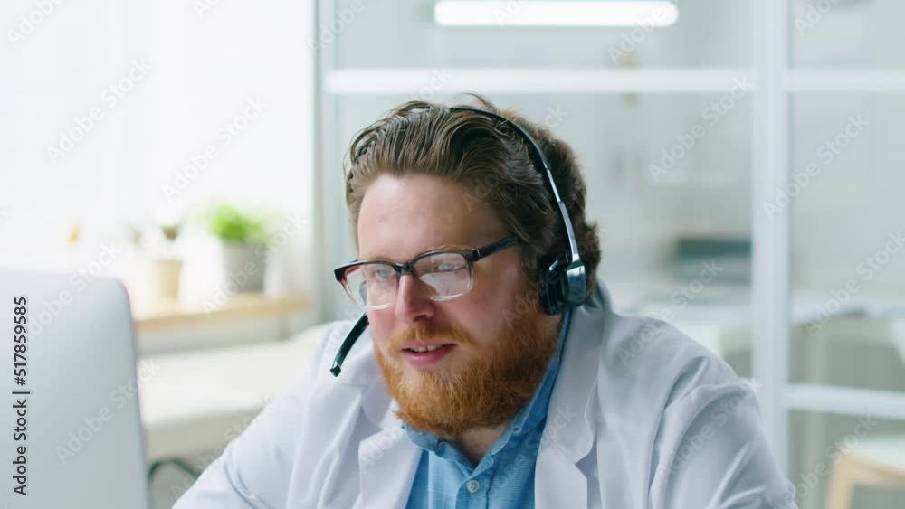Bearded male doctor in headset explaining how to do self checkup via online video call on computer while working remotely from medical office Bearded male doctor in headset explaining how to do self checkup via online video call on computer while working remotely from medical office