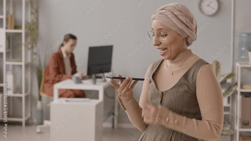 Medium slowmo of enthusiastic young Muslim woman recording voice message on smartphone at workplace in office while her female colleague working on computer in blurred background Medium slowmo of enthusiastic young Muslim woman recording voice message on smartphone at workplace in office while her female colleague working on computer in blurred background