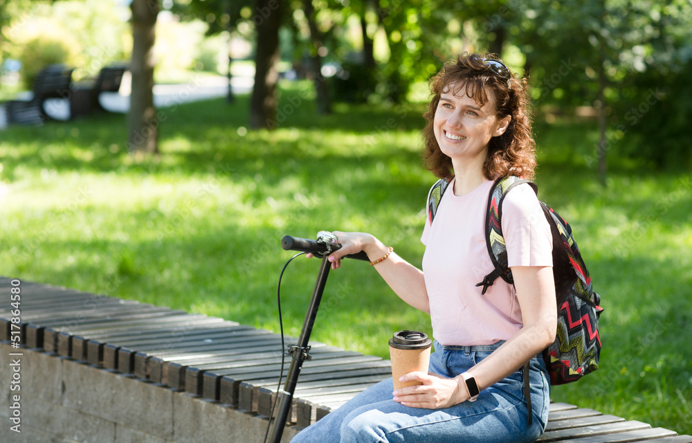 middle aged woman with a backpack and scooter rests in the park and drinks coffee on a summer day middle aged woman with a backpack and scooter rests in the park and drinks coffee on a summer day