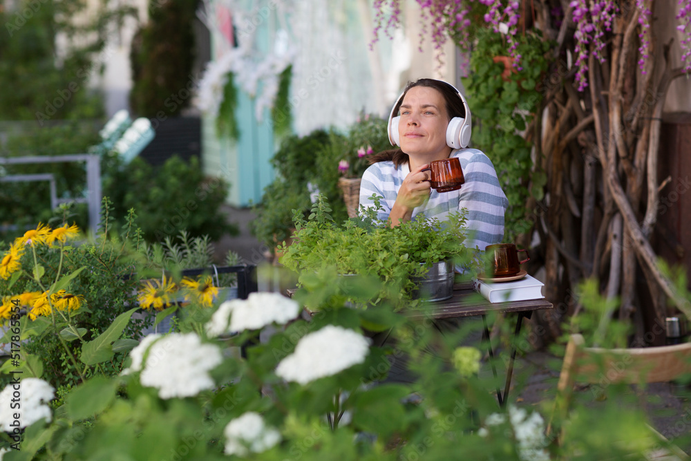 middle aged woman wearing headphones with a cup of tea relaxed in the garden near the house in summer. concept without stress. mental health. Slow life. Enjoying the little things. middle aged woman wearing headphones with a cup of tea relaxed in the garden near the house in summer. concept without stress. mental health. Slow life. Enjoying the little things.