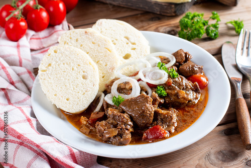 Pork goulash meat with dumplings on white plate, cutlery, garlic, onion, tablecloth in the background - typical Czech food