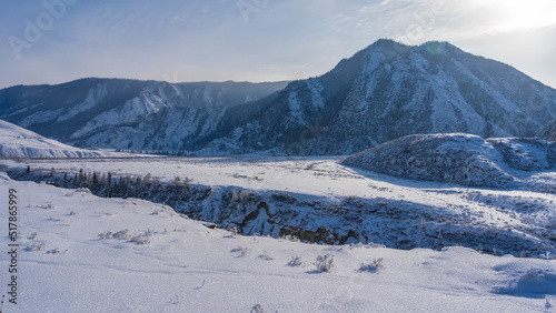 The gorge crosses a snow-covered valley. Trees are visible on steep slopes. Pure white snow lies on the ground. A picturesque mountain range against the sky. Altai