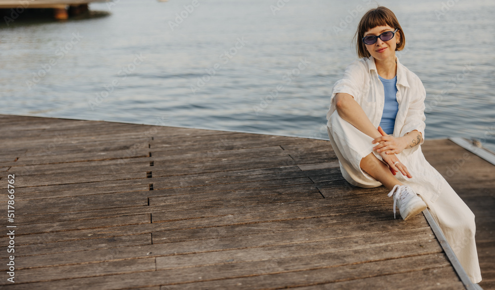 Pretty young brown-haired caucasian lady spends weekend outdoors sitting by sea. Girl with bob haircut wears sunglasses, shirt and pants. Relaxation concept