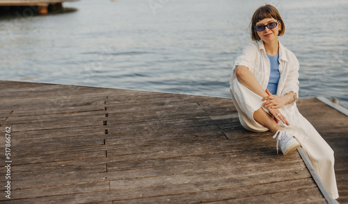 Pretty young brown-haired caucasian lady spends weekend outdoors sitting by sea. Girl with bob haircut wears sunglasses, shirt and pants. Relaxation concept