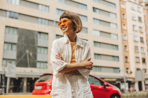 Smiling young caucasian girl looking at camera spending time outdoors during day. Brown-haired woman with bob haircut wears sunglasses, white shirt and pants. Summer vacation concept. #517868738