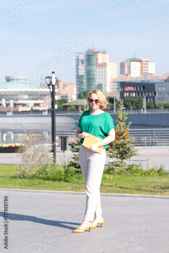 A happy young blonde student girl in sunglasses walking along a city street, carries an orange digital tablet. Cute smiling hipster student girl on the background of office buildings and blue sky