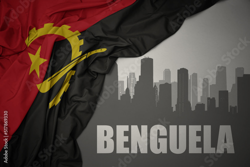 abstract silhouette of the city with text Benguela near waving colorful national flag of angola on a gray background. photo
