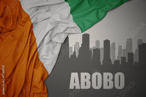 abstract silhouette of the city with text Abobo near waving colorful national flag of cote divoire on a gray background.