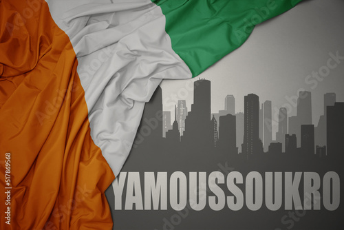 abstract silhouette of the city with text Yamoussoukro near waving colorful national flag of cote divoire on a gray background. photo