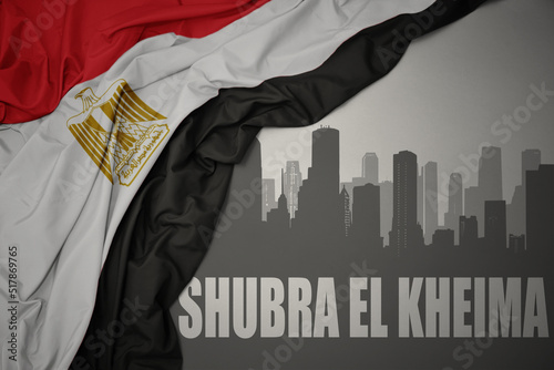 abstract silhouette of the city with text Shubra El Kheima near waving colorful national flag of egypt on a gray background. photo