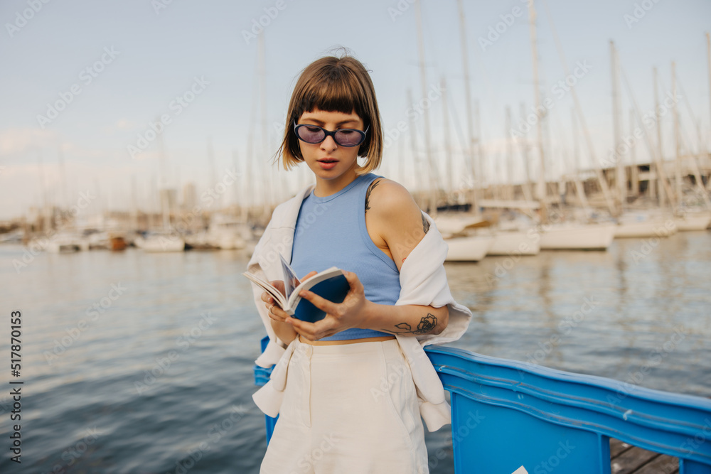 Beautiful young caucasian girl leafing through book spending time outdoors in summer. Brown-haired woman with bob haircut wears sunglasses, top and pants. Stylish vacation concept