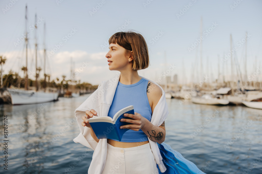 Young caucasian woman with bob haircut looks away relaxing near sea in yacht club summer. Model wears tank top, sweatshirt and pants. Lifestyle, leisure concept