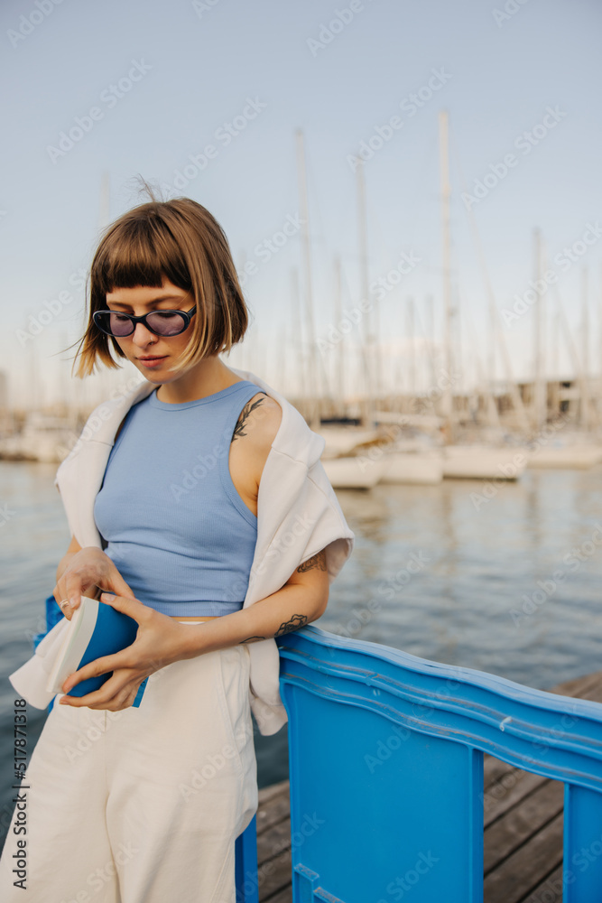 Young caucasian woman in sunglasses holding book standing outdoors yacht club. Brown-haired with bob haircut wears T-shirt, sweatshirt and pants summer. Rest time concept.