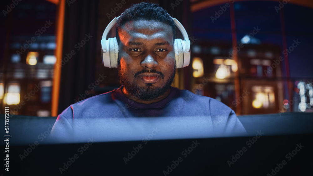 Obraz premium Young Handsome Black Man in Headphones Working from Home on Laptop Computer in Cozy Stylish Loft Apartment. Creative Male Smiling, Checking Social Media, Browsing Internet. Flat with Big Windows.