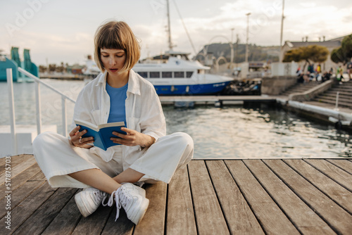 Cute young caucasian woman enthusiastically reads book sitting on pier of sea station. Brown-haired with bob haircut wears casual clothes. Lifestyle concept