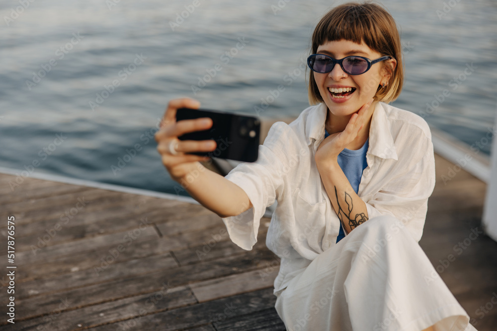 Cheerful young caucasian girl in sunglasses uses smartphone sitting by sea evening. Brown-haired woman with bob haircut wears casual clothes spring. Concept of modern devices