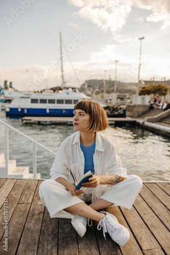 Calm young caucasian girl with book in her hands looks away sitting outdoors near sea. Brown-haired woman bob haircut wears casual spring clothes. People and free time concept