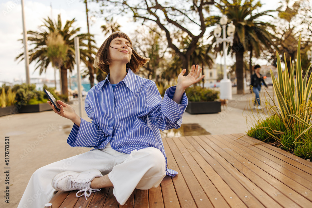 Satisfied young caucasian girl with phone closing her eyes is resting sitting on bench in springtime. Brown-haired woman with bob haircut wears casual clothes. Positive emotions concept