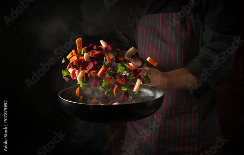 Canvas Print The hand of a professional chef throws pieces of vegetables into a hot frying pan with steam on a black background