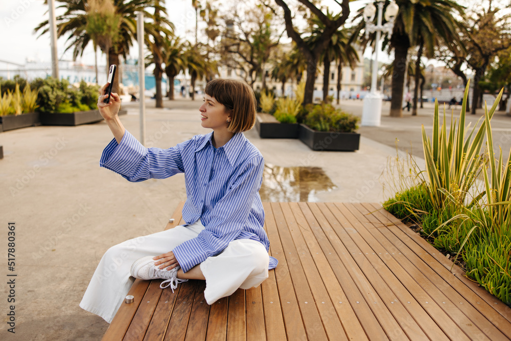 Cute young caucasian girl takes selfie on phone sitting bench in European city. Brown-haired woman with bob haircut wears casual clothes. Technology concept Cute young caucasian girl takes selfie on phone sitting bench in European city. Brown-haired woman with bob haircut wears casual clothes. Technology concept