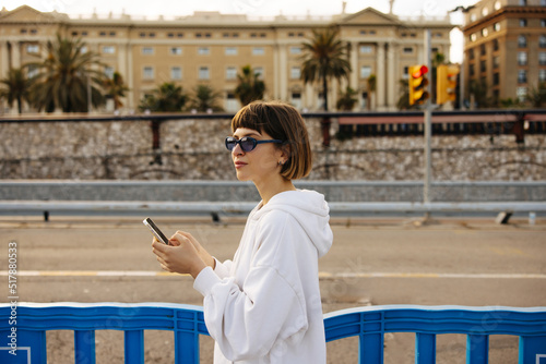 Nice young caucasian woman typing sms on smartphone relaxing outdoors during daytime. Brown haired with bob haircut wears white hoodie, sunglasses in spring. City life concept © Look!