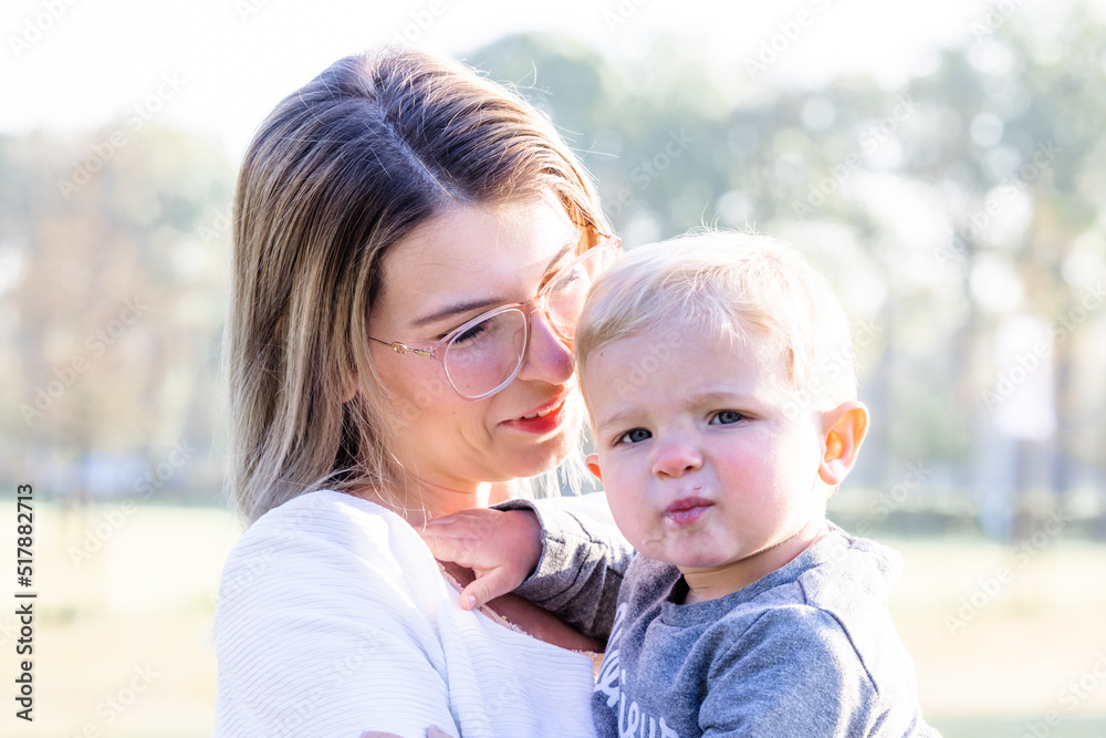 Happy young mother with a small child or toddler in her arms, hugging and cuddling, standing in the middle of a park in the rays of the rising morning sun. High quality photo Happy young mother with a small child or toddler in her arms, hugging and cuddling, standing in the middle of a park in the rays of the rising morning sun. High quality photo