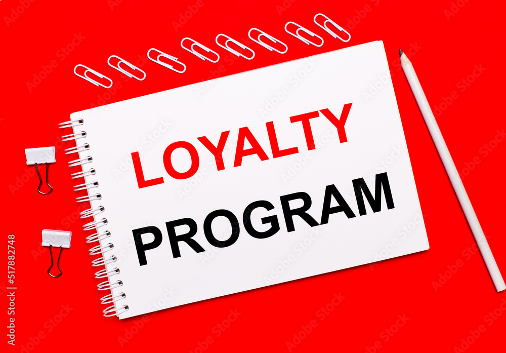 On a bright red background, a white pencil, white paper clips, and a white notebook with the text LOYALTY PROGRAM