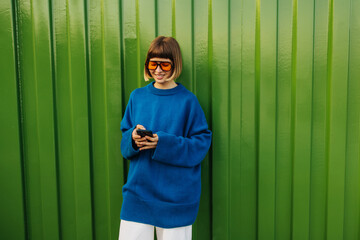 Positive young caucasian woman uses modern smartphone standing against green wall. Brown-haired with bob haircut wears sunglasses, blue sweater. Technology concept