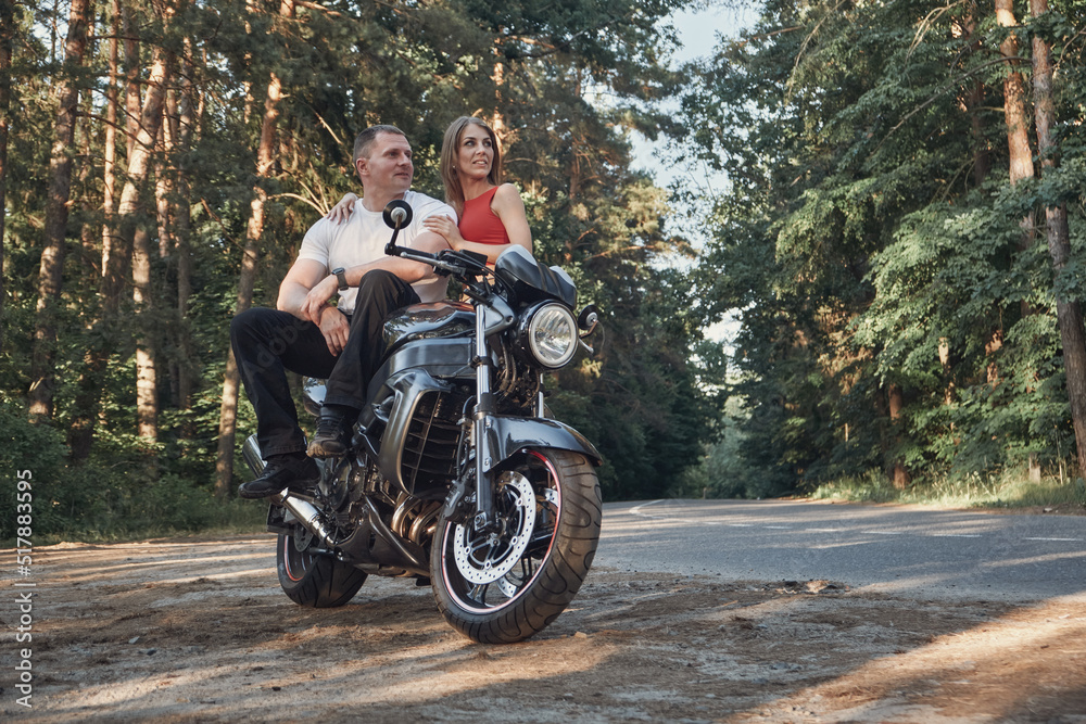 Young beautiful couple talking and having fun, sitting on a motorcycle, traveling together on a forest road