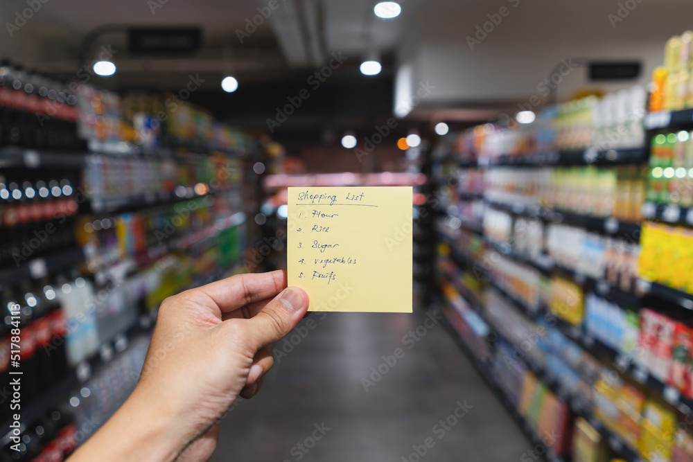 Hand holding a yellow note with hand writing shopping list in blur supermarket aisle background